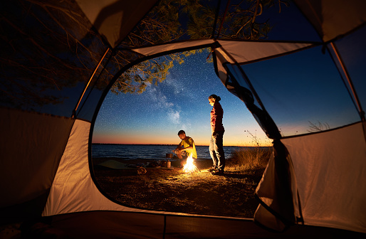 Camping on lake shore at sunset, view from inside tourist tent. Young romantic couple, man and woman preparing dinner on campfire on blue sea water background. Tourism, recreation and love concept.