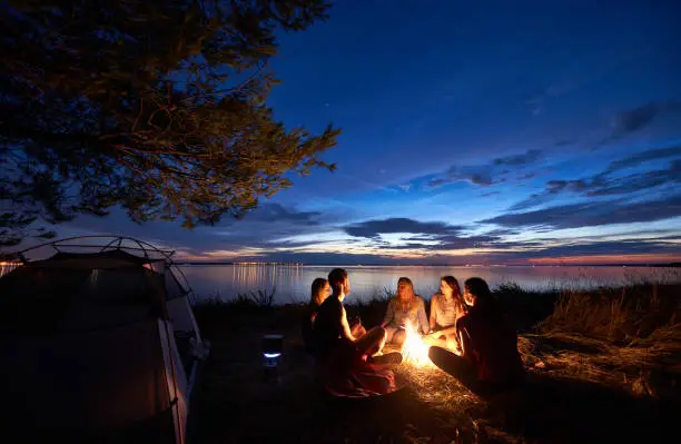 Night summer camping on sea shore. Group of five young tourists sitting on the beach around campfire near tent under beautiful blue evening sky. Tourism, friendship and beauty of nature concept.