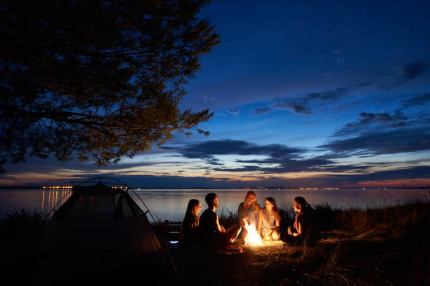夜夏に海岸にキャンプします。夕方の空の下でテント近くのキャンプファイヤーの周りの若い観光客のグループ - friendship camping night campfire ストックフォトと画像