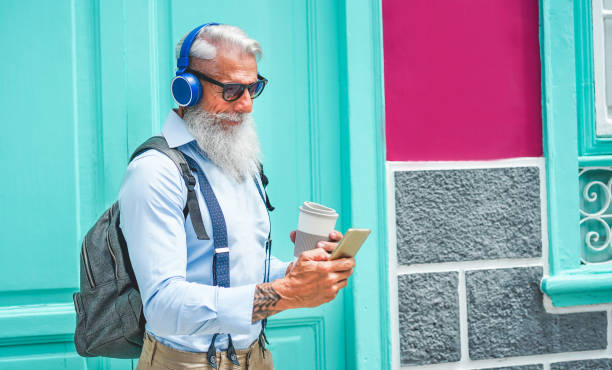 時髦的資深男子使用音樂智慧手機應用程式和飲用咖啡市中心中心戶外-成熟的時尚男性與新的趨勢技術和快樂的老年人生活方式概念的樂趣 - 態度瀟灑 圖片 個照片及圖片檔