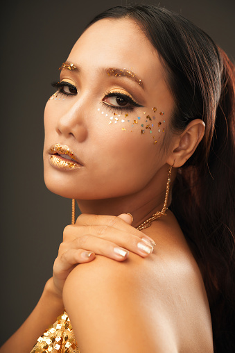 Side view of gorgeous young ethnic woman in festive clothes with sparkling golden glitter on lips, eyebrows and cheekbones holding hand on shoulder and looking at camera over grey studio background