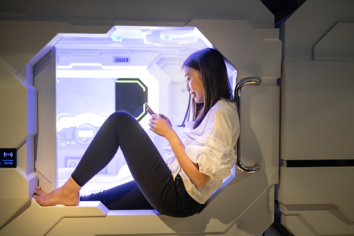 an asian chinese female staying in a capsule hotel room sitting at the edge of the door looking at her mobile phone surfing internet