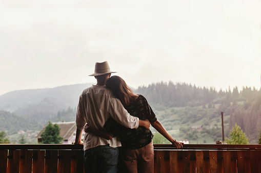 hipster couple hugging on porch of wooden house looking at mountains in evening sunset, romantic moment, summer relaxing together concept, space for text