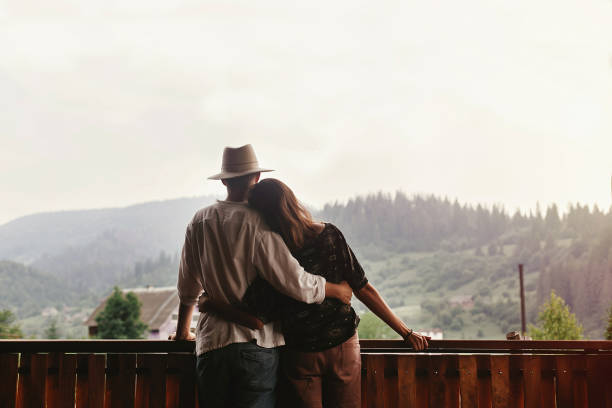 hipster couple hugging on porch of wooden house looking at mountains in evening sunset, romantic moment, summer relaxing together concept, space for text hipster couple hugging on porch of wooden house looking at mountains in evening sunset, romantic moment, summer relaxing together concept, space for text log cabins stock pictures, royalty-free photos & images