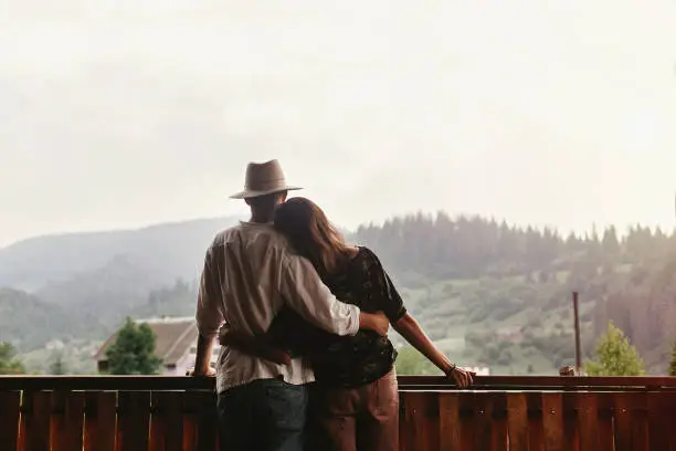 Photo of hipster couple hugging on porch of wooden house looking at mountains in evening sunset, romantic moment, summer relaxing together concept, space for text