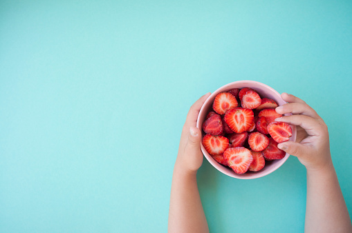 Child's hands holding a pink bowl full of beautiful fresh juicy strawberries on pastel blue background with text space.