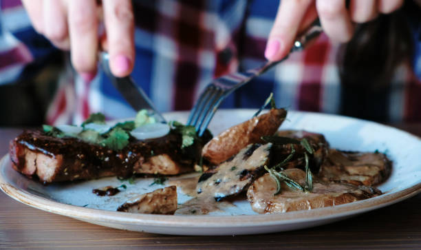 closeup woman's hand cutting piece of pork cooked on the coals with potatoes, gravy and greens on plate and eating. - hot couture imagens e fotografias de stock