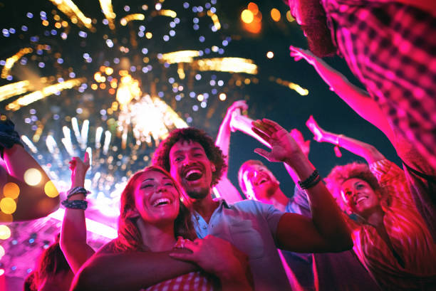 Friends celebrating on a night out. Closeup low angle view of group of multi ethnic young adults dancing and enjoying an open air concert on a summer afternoon. They are standing in a circle while fireworks popping in the background. public celebratory event stock pictures, royalty-free photos & images