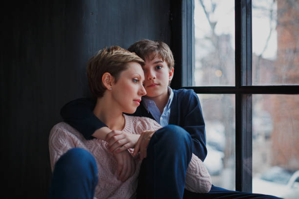 A teenage boy hugs his young mother with short hair and looks at the camera with sad eyes. stock photo