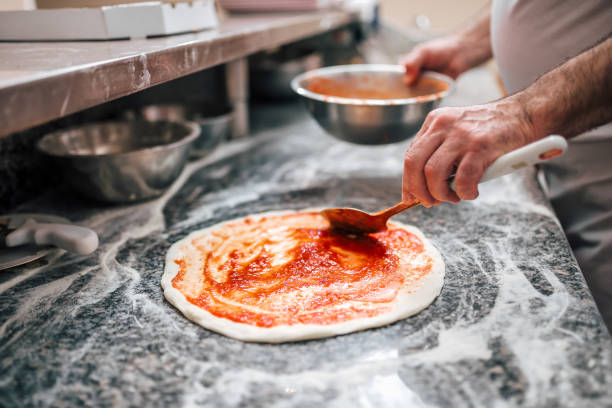 istock Raw pizza, preparation in traditional style. Adding tomato sauce on a pizza dough. 1002939968