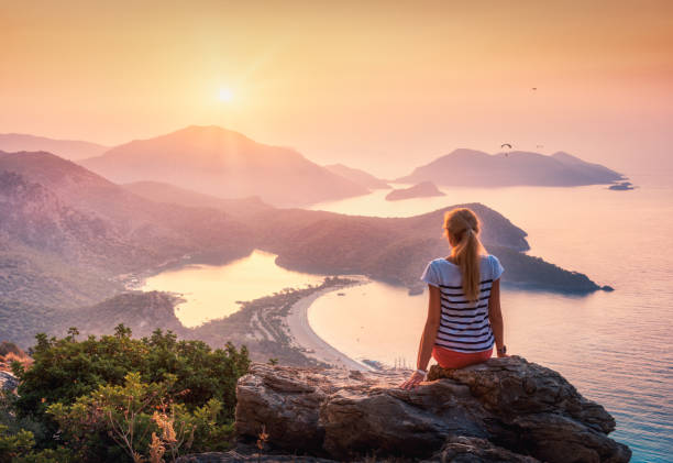 jeune femme assise sur le dessus de la roche et en regardant sur le bord de mer et les montagnes coloré coucher de soleil en été. paysage avec la fille, mer, montagnes et ciel orange avec le soleil. oludeniz, turquie. - climbing rock climbing rock mountain climbing photos et images de collection