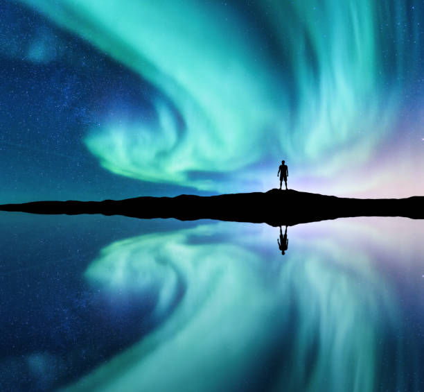 Photo of Northern lights and silhouette of standing man in the hill in Norway. Aurora borealis and man. Stars and green polar lights. Night landscape with aurora, lake, sky reflection in water. Travel. Concept