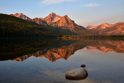 Sunrise lights up McGown Peak and the Sawtooth range as the calm waters of Stanley Lake reflect this stunning Idaho nature scene.