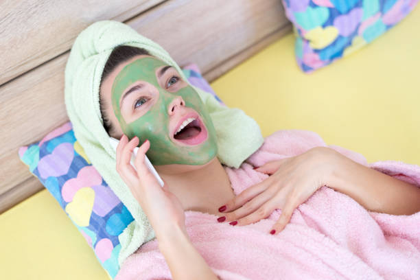 Woman with facial mask gossiping over the smart phone at home while lying in the bed.