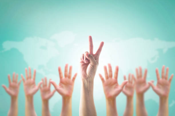 istock Leader's two fingers victory sign among blur hands crowd group: Many people blurred palms raising up upward on vintage blue sky background map: World participation, leadership volunteer concept: V day 1003209808