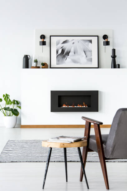istock Table next to grey armchair in white loft interior with poster above black fireplace. Real photo 1003209972