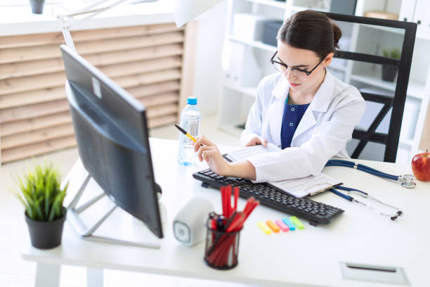 une belle jeune fille dans une robe blanche est assis à un bureau d’ordinateur avec des documents et un stylo dans ses mains. - patient hospital doctor nurse photos et images de collection