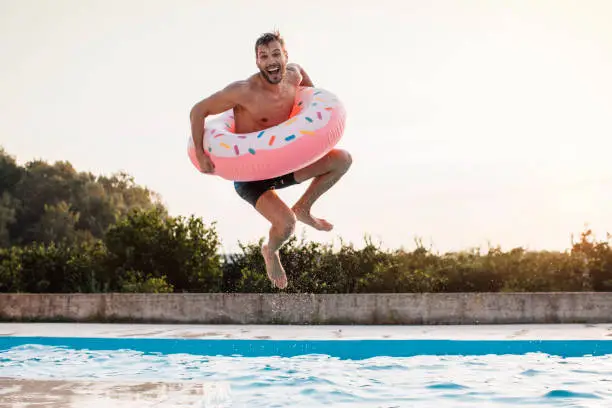 Young man enjoying time at the swimming pool jumping into the water with inflatable ring