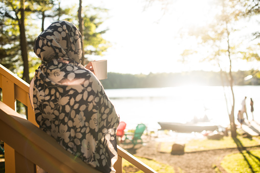 Young professional Muslim woman wearing Hijab on vacation enjoying morning coffee on a lake house deck over looking the water