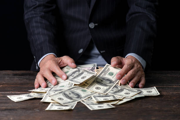 Pile of paper currency . Man hands in pinstripe suit sweeping pile of US dollar banknotes on old wooden table and black background,business concept. greedy corporation stock pictures, royalty-free photos & images