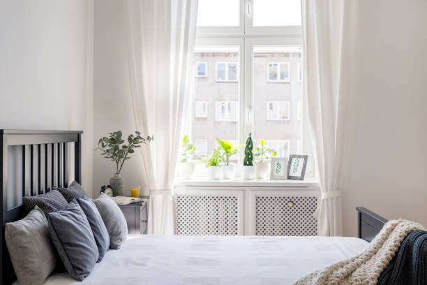 istock Knit blanket and pillows on bed in white hotel bedroom interior with drapes at window. Real photo 1003384620