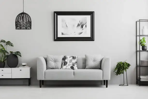 Black industrial bookcase and a plant stand next to an upholstered sofa in a gray living room interior with place for a coffee table. Real photo