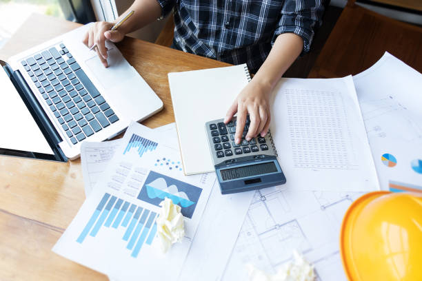 istock Asian engineer , architect woman using calculator for accounting and analyzing building structure from blueprint and paper chart in front of computer laptop at office work space. 1003408944