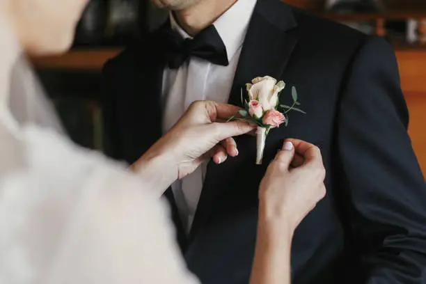 beautiful bride putting on stylish simple boutonniere with roses on groom black suit. wedding morning preparations