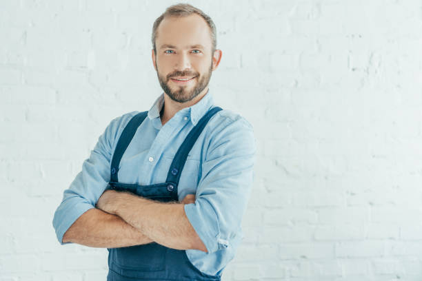 istock smiling bearded worker in overalls posing with crossed arms 1003472512