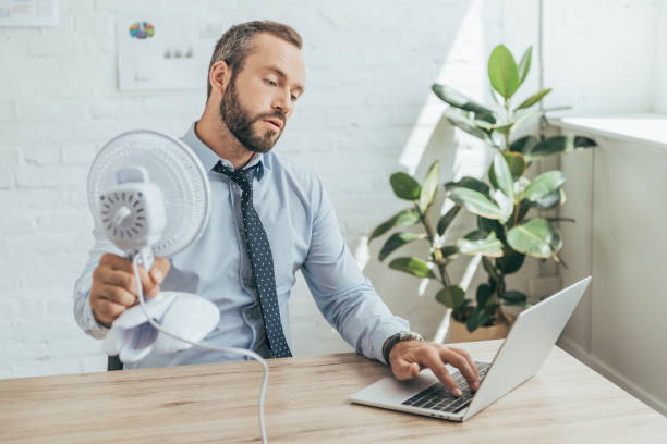 бизнесмен охлаждения себя с электрическим вентилятором при использовании ноутбука в офисе - office fan стоковые фото и изображения