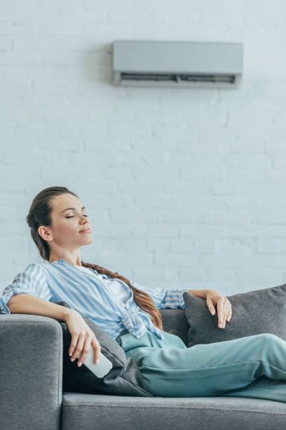 istock woman resting on sofa with air conditioner on wall, summer heat concept 1003476324