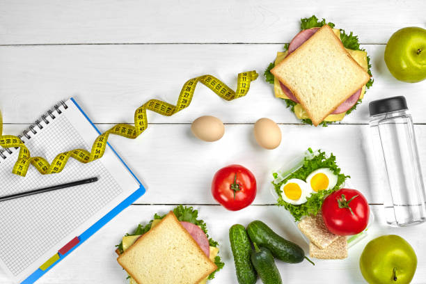 istock Top view planning notebook with copy space and healthy breakfast. Sandwich with ham and cheese, eggs, cucumber, tomato and bottle of water on white wooden table 1003479946