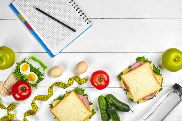 istock Top view planning notebook with copy space and healthy breakfast. Sandwich with ham and cheese, eggs, cucumber, tomato and bottle of water on white wooden table 1003480512