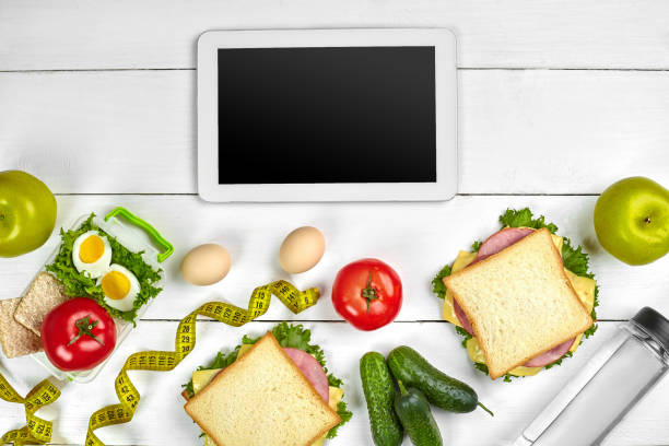 istock White tablet computer with a blank screen on the kitchen table with sandwiches, fresh vegetables, bottle of water and apple. Lunch 1003480808