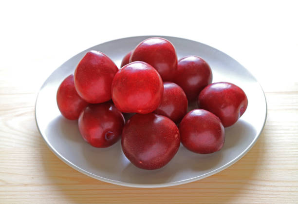 istock Fresh Gulf Ruby Plum Fruits Piled up on White Plate Served on Wooden Table 1003481396