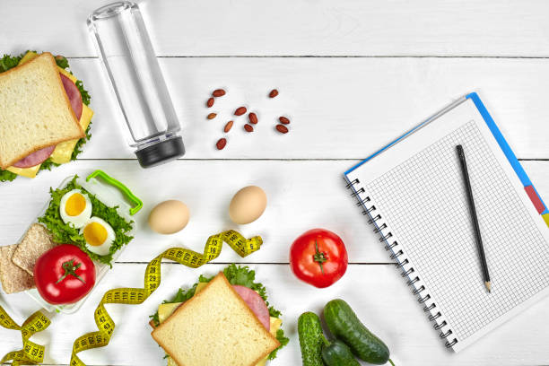 istock Top view planning notebook with copy space and healthy breakfast. Sandwich with ham and cheese, eggs, cucumber, tomato and bottle of water on white wooden table 1003481488