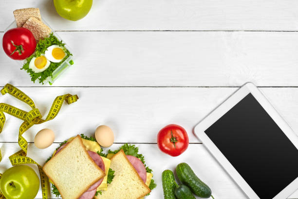 istock White tablet computer with a blank screen on the kitchen table with sandwiches, fresh vegetables, bottle of water and apple. Lunch 1003484106