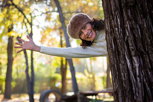 Smiling middle aged woman comes out behind tree trunk with arm extended in the park during fall season. Female model, autumn fashion sales concept