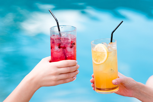 cropped shot of women clinking glasses of delicious cocktails in front of swimming pool