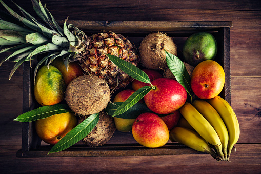 Wooden crates with assorted tropical fruits in rustic kitchen. Natural lighting. Mangos, pineapples, coconuts, bananas