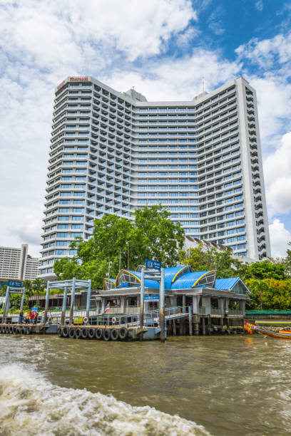 Bangkok, Thailand - May 02 2018:  The Chao Phraya river runs through the centre of Thailand’s capital city, Bangkok.  The express boat service is part of the cities major transport network, with multiple Piers along the route.  Here we see the Si Phraya Pier, with the towering Royal Orchid Sheraton hotel in the background.