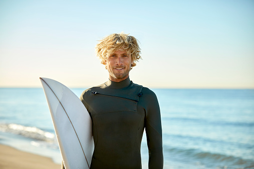 Portrait of confident mid adult man carrying surfboard at beach. Smiling male surfer is wearing wetsuit against sea. He is on summer vacation.