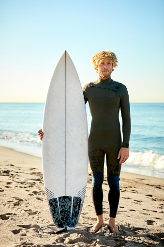 Full length portrait of confident male surfer with surfboard at beach. Handsome man is wearing wetsuit against sky. He is on summer vacation.