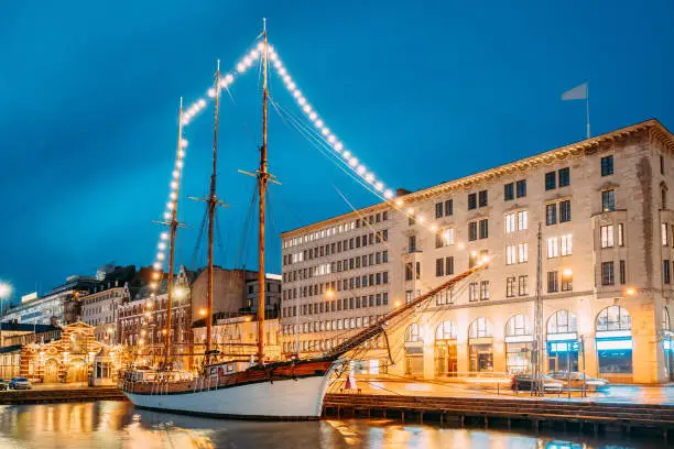 Photo of Helsinki, Finland. Old Wooden Sailing Vessel Ship Schooner Is Moored To The City Pier, Jetty. Unusual Cafe Restaurant In City Center In Evening Or Night Illumination