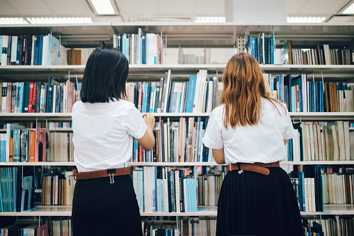 Two young Asian students talking after classes in Bangkok, Thailand. They're wearing traditional Thai school uniforms, searching for books in the campus library.