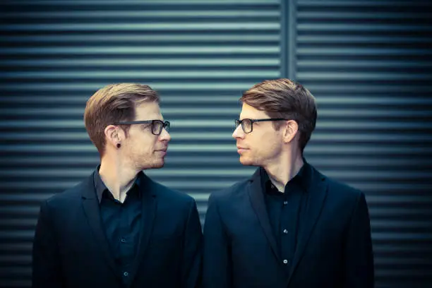 twin brothers with black suits and glasses standing and looking into each other eyes.