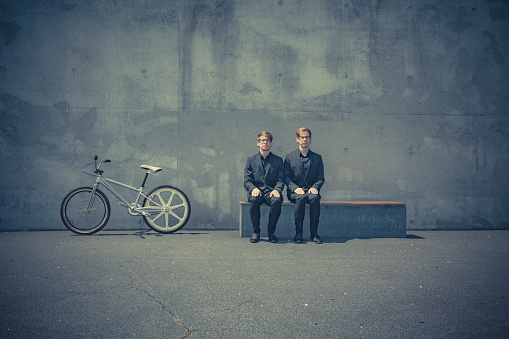 twin brothers with black suits and glasses sitting on a bench with a bmx bicycle next to them.
