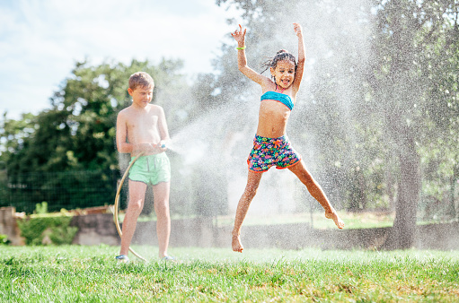 Hot summer days activity: happy little girl jumps under water, when brother pours her from garden hose