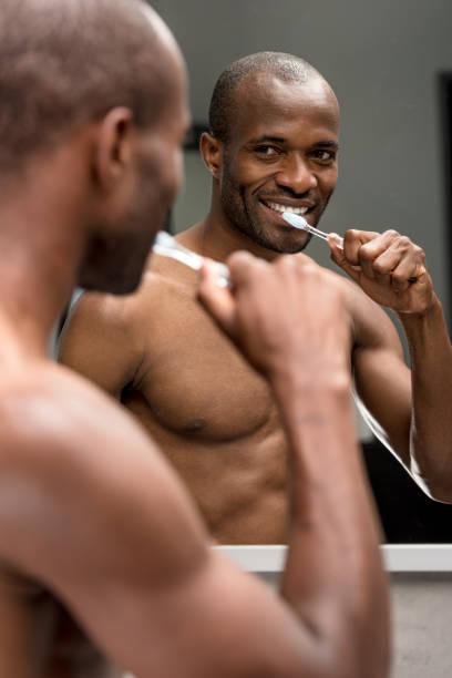 istock smiling shirtless african american man brushing teeth and looking at mirror 1003751706