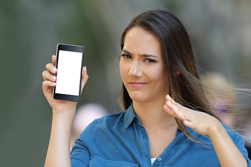 Doubtful woman showing a blank phone screen and looking at camera on the street